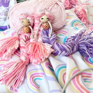 May include: Two handmade mermaid dolls with wooden bead heads, yarn hair, and tassel tails. One doll has pink and white yarn, the other has purple and white. They are decorated with small silver crowns and sit on a rainbow-patterned fabric.