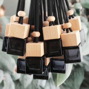 Puede incluir: Un conjunto de ambientadores para coche de cristal negro con tapas de madera natural y cordones negros. Los ambientadores tienen forma cuadrada y están colgados juntos. El fondo es borroso con follaje verde.