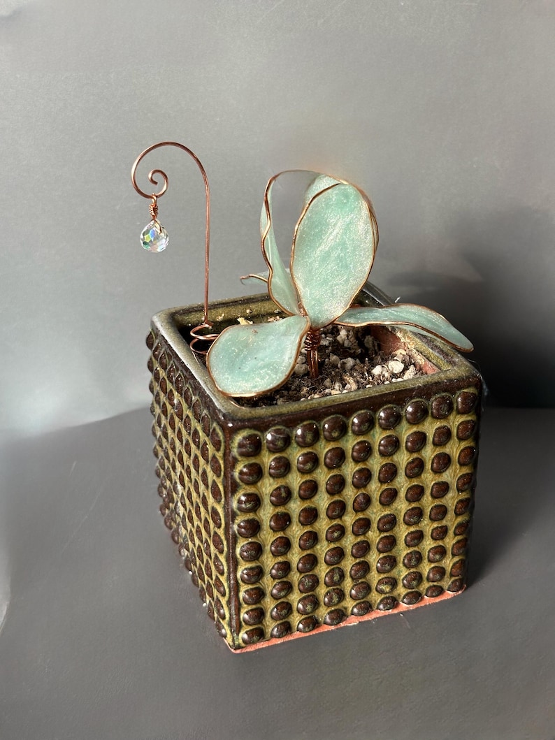 Op de afbeelding: Een vierkante keramische plantenbak met een gestructureerd, gestippeld patroon in bruin en groen. Het bevat een vetplant met lichtgroene bladeren. Een koperdraad accent met een kristallen kraal voegt een decoratief tintje toe.