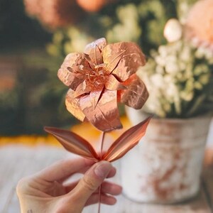 以下が含まれることがあります： 詳細な花びらと葉を持つ銅色の金属製の花。 手で持たれており、その複雑なデザインと金属的な輝きを示しています。 茎と葉は細く、手作りの花の芸術の繊細な外観を加えています。