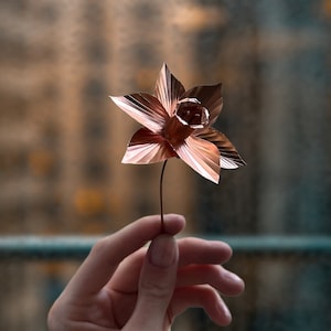 Può includere: Un fiore di narciso in metallo color rame con un design dettagliato dei petali. Il fiore è tenuto da una mano, mostrando l'intricata maestria artigianale. Lo stelo è sottile e simile a un filo, aggiungendo all'estetica delicata. Lo sfondo è sfocato.