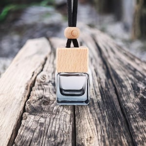 May include: A clear, square glass bottle with a light wood cap and a black cord for hanging. The bottle is filled with a clear liquid. The background is a blurred wooden surface.