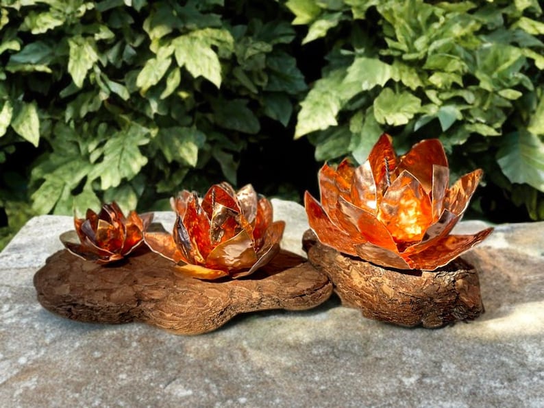 May include: Three copper lotus flower sculptures of varying sizes sit on a piece of driftwood. The flowers have a warm, metallic orange hue, and the driftwood is a rich brown. Green foliage forms the background.