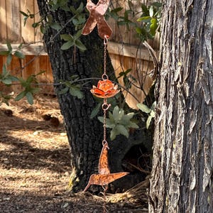 May include: A copper-colored rain chain with hummingbird and lotus flower designs. The chain is suspended from a tree branch and features intricate cut-out details. The chain is made of metal and is designed for outdoor use.
