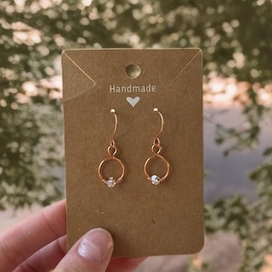 May include: Handmade copper wire earrings with a small, clear crystal bead in the center of each hoop. The earrings are displayed on a brown card with the word "Handmade" and a heart symbol.