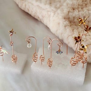 Può includere: Una collezione di espositori di gioielli artigianali in filo di rame. Gli espositori presentano vari design, tra cui una libellula, una stella e composizioni floreali. Ogni espositore ha una base a spirale e contiene orecchini delicati con accenti di cristallo e fiori.