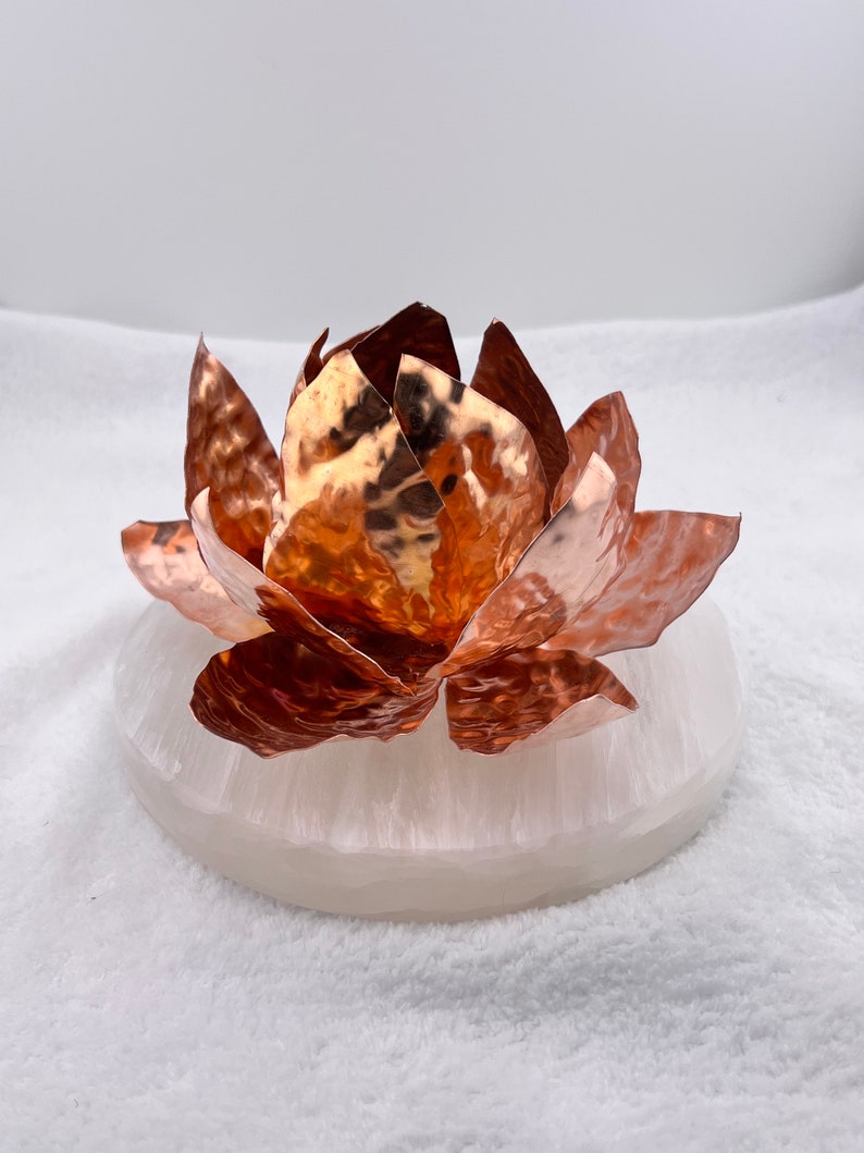 May include: A copper lotus flower sculpture with textured petals sits atop a round, white, translucent base. The copper petals have a reflective, metallic finish. The base appears to be made of a smooth, solid material.