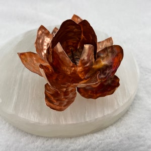 May include: A handcrafted copper lotus flower sculpture with detailed petals. The flower is placed on a white, round, selenite crystal base. The copper has a rich, warm brown color.