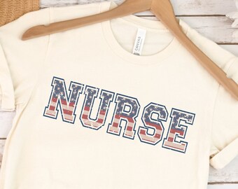 Puede incluir: Una camiseta blanca con la palabra "NURSE" impresa en letras azules con un patrón de bandera estadounidense roja, blanca y azul.