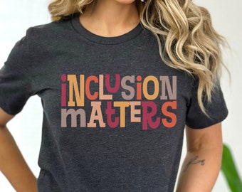以下が含まれることがあります： カラフルな文字で「INCLUSION MATTERS」と書かれたダークヘザーグレーのTシャツです。