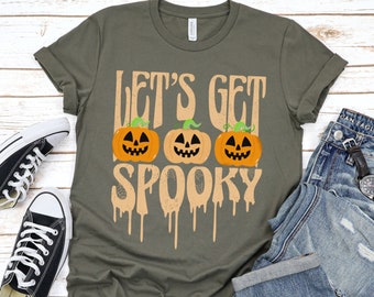 Op de afbeelding: Een groen T-shirt met de tekst "Let's Get Spooky" in oranje en een druppelend effect. Drie oranje pompoenen met zwarte ogen en monden bevinden zich boven de tekst.
