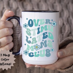 May include: White ceramic mug with a blue rim and handle. The mug has a graphic design that says "Over Stimulated Moms Club" in a retro font with blue, green, and yellow colors. The design also includes small flower accents.