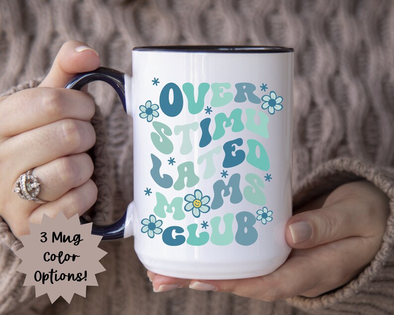 May include: White ceramic mug with a blue rim and handle. The mug has a graphic design that says "Over Stimulated Moms Club" in a retro font with blue, green, and yellow colors. The design also includes small flower accents.
