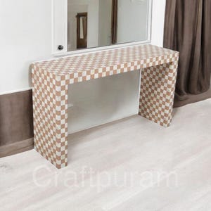 Könnte beinhalten: Ein rechteckiger Konsolentisch mit einem braun-weißen Schachbrettmuster. Der Tisch hat ein schlichtes, offenes Design mit zwei Beinen und einer langen Tischplatte. Der Tisch steht vor einer weißen Wand mit einem Spiegel darüber.