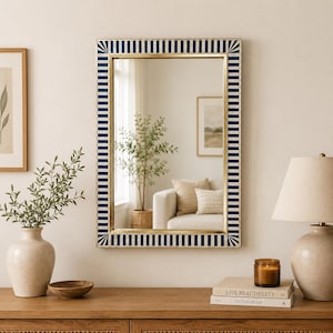May include: Rectangular wall mirror with a gold frame and a navy blue and white striped border. The mirror reflects a room with a white sofa, a plant, and a lamp. The mirror is a decorative home decor item.