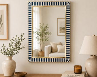 May include: Rectangular wall mirror with a gold frame and a navy blue and white striped border. The mirror reflects a room with a white sofa, a plant, and a lamp. The mirror is a decorative home decor item.