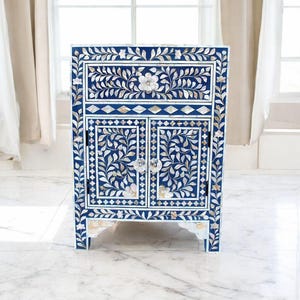 May include: A navy blue cabinet with a floral design. The cabinet has two doors and a drawer, all adorned with a white floral inlay. The cabinet has clear crystal knobs and is set against a white marble floor.
