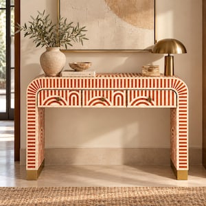 Op de afbeelding: Een rechthoekige sidetable met een herhalend patroon van oranje en crèmekleurige gestreepte bogen. De tafel heeft drie lades met kleine ronde messing knoppen en rust op twee massieve poten met gouden metalen accenten.