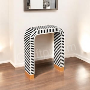 Könnte beinhalten: Ein moderner Konsolentisch mit einem markanten Design. Der Tisch hat ein schwarz-weiß gestreiftes Muster auf der Oberseite und den Seiten, mit einer geschwungenen, U-förmigen Silhouette. Die Basis der Beine hat eine goldene Oberfläche. Der Tisch ist etwa 76 cm hoch.