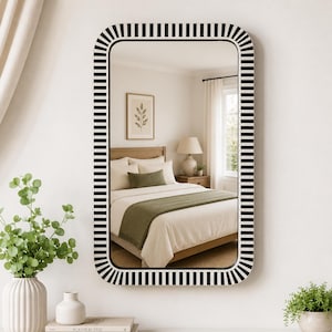 May include: Rectangular mirror with a black and white striped frame. The mirror reflects a bedroom scene with a bed, lamp, and artwork. The frame has rounded corners and a modern design.