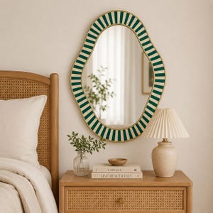 May include: An oval-shaped wall mirror with a gold frame and a green and white striped border. The mirror is hanging above a wooden nightstand with a lamp, books, and a small vase. The nightstand has a woven design.