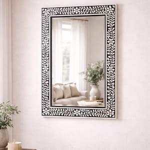 May include: Rectangular mirror with a black frame adorned with a white floral pattern. The mirror reflects a room with a sofa, vase, and plants, creating a decorative and functional home decor piece.