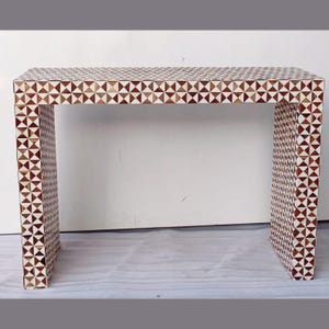 Puede incluir: Una consola rectangular con un patrón geométrico. La mesa está hecha de un material de color claro, posiblemente madera, y está cubierta con un patrón repetitivo de triángulos marrones y blancos. La mesa tiene un diseño simple y moderno.
