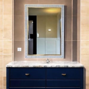 May include: A rectangular mirror with a striped white and light blue frame hangs above a navy blue vanity with gold-coloured handles. The vanity has a marble countertop and a chrome tap. The background features beige tiles.
