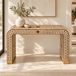 May include: A rectangular console table with a waterfall edge design, featuring an intricate brown and white floral and leaf inlay pattern. The table has a smooth top surface and stands on two wide, continuous side panels with gold-toned feet.