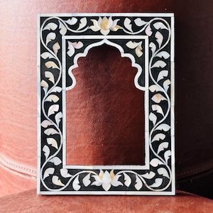 May include: Rectangular black and white decorative frame with an arched top. The frame is inlaid with mother-of-pearl and features floral and leaf designs. The frame is likely for a photograph or artwork.
