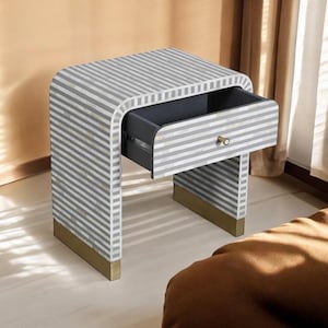 Puede incluir: Una mesita de noche moderna con un patrón de rayas grises y blancas. El cajón está abierto, revelando un interior oscuro. La base tiene detalles dorados. La mesita mide aproximadamente 61 cm de alto, 51 cm de ancho y 41 cm de profundidad.