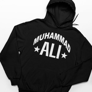 May include: Black hooded sweatshirt with the name "MUHAMMAD ALI" in white, arched across the chest. Two white stars flank the name. The sweatshirt has a front pocket and a drawstring hood.