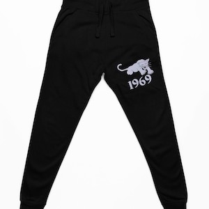 May include: Black sweatpants with a drawstring waist and elastic cuffs. A light gray panther graphic and the number "1969" are printed on the left leg. The pants are made of a soft, comfortable material.