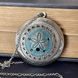 May include: Silver-toned locket necklace with a turquoise and copper sand dollar design. The round pendant features intricate filigree detailing around the edges and a delicate chain. A vintage-inspired piece.