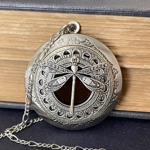 Peut inclure: Collier avec médaillon en argent orné d'un motif de libellule. Le médaillon a une fenêtre transparente pour révéler une photo ou un autre petit objet. La chaîne est en argent et a un maillon délicat.