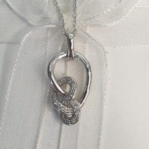 May include: A silver-coloured necklace featuring a pendant with a polished teardrop-shaped outer frame and an inner interlocking knot design. The knot element is encrusted with small, clear, sparkling stones that catch the light, suspended from a delicate chain.