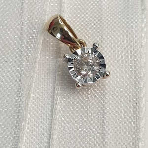 May include: A gold pendant featuring a round, clear, faceted stone set in a silver-toned, sunburst-patterned metal frame. The pendant includes a polished gold bail at the top, designed for attachment to a chain, showcasing a two-tone metal aesthetic.
