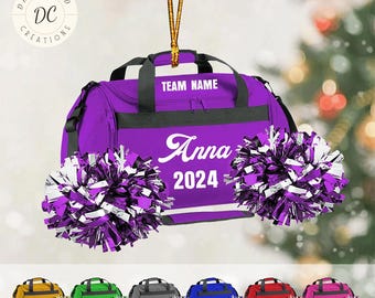 May include: A purple duffel bag ornament with the text "TEAM NAME", "Anna", and "2024". The ornament has black accents and is paired with purple and silver pom-poms. The bottom row shows similar ornaments in various colors.