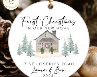 May include: A white ceramic Christmas ornament with a watercolor illustration of a house, pine trees, and falling snow. Text reads "First Christmas in Our New Home," address, names, and the year 2024. The ornament has a jute string for hanging.