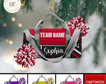 May include: A red, black, and gray duffel bag ornament with the text "TEAM NAME" and "Sophia". The ornament has a gray shoulder strap and handle. The image also shows red and white pom-poms and other color variations of the bag.