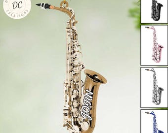 May include: A gold-colored saxophone ornament with the name "JOHN" on the bell. The ornament is hanging from a green branch. The words "PERSONALIZED DESIGN" are at the bottom. Additional saxophone designs are shown on the right.