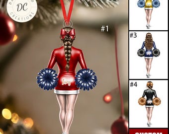 May include: A Christmas ornament featuring a cheerleader in a red uniform with a white skirt, holding navy blue pom-poms. The ornament has a red ribbon for hanging and the name "Lucy" is on the skirt. Additional cheerleader designs are shown.