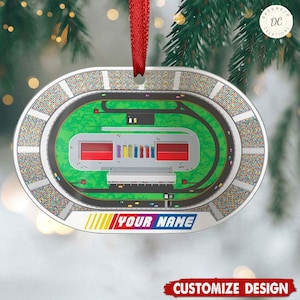 Peut inclure: Une décoration de Noël ovale blanche avec une illustration colorée d'un circuit de course. La piste est entourée d'une foule de personnes. Le texte "YOUR NAME" est imprimé en rouge, bleu et jaune au bas de la décoration.