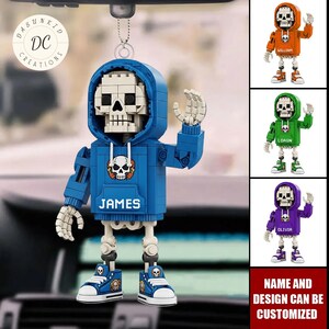 May include: A blue skeleton figure wearing a hoodie and sneakers, with the name "JAMES" on the front. The figure is hanging from a car's rearview mirror. Additional figures in orange, green, and purple hoodies are shown. Text reads "NAME AND DESIGN CAN BE CUSTOMIZED."