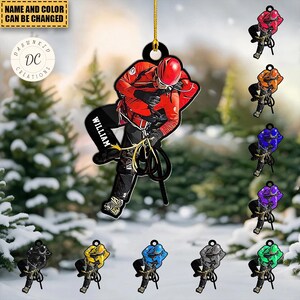 May include: A personalized ornament featuring a climber in a red suit and helmet, with the name "WILLIAM" on the leg. The ornament is suspended by a gold string. Additional color variations are displayed, including orange, blue, purple, and green.