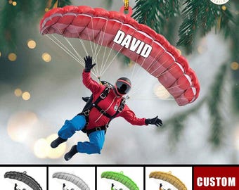 Può includere: Un paracadute rosso e bianco con il nome "DAVID" stampato su di esso. Un paracadutista è attaccato al paracadute e indossa una tuta rossa, un casco nero e guanti neri. Il paracadute è appeso a un filo dorato, il che suggerisce che sia un ornamento natalizio.