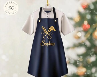May include: A navy blue apron with gold scissors, a comb, and a hairdryer design, with the name "Sophia" in gold script. The apron hangs on a white shirt. The text "Customize Your Design" is at the top.