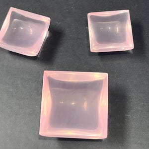 May include: Three pink, square-shaped, crystal dishes with a slightly curved, concave interior.