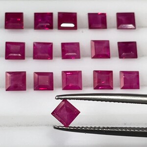 May include: A collection of 16 square-shaped ruby gemstones, arranged in rows on a white surface. One ruby is being held by tweezers.