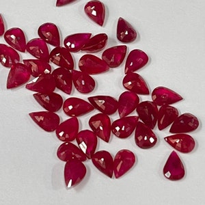 May include: A collection of pear-shaped ruby gemstones, with a deep red color, scattered on a white surface.